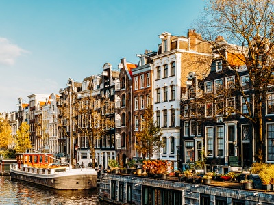 Een gracht in Amsterdam met traditionele grachtenpanden, een woonboot en herfstbomen verlicht door de zon.