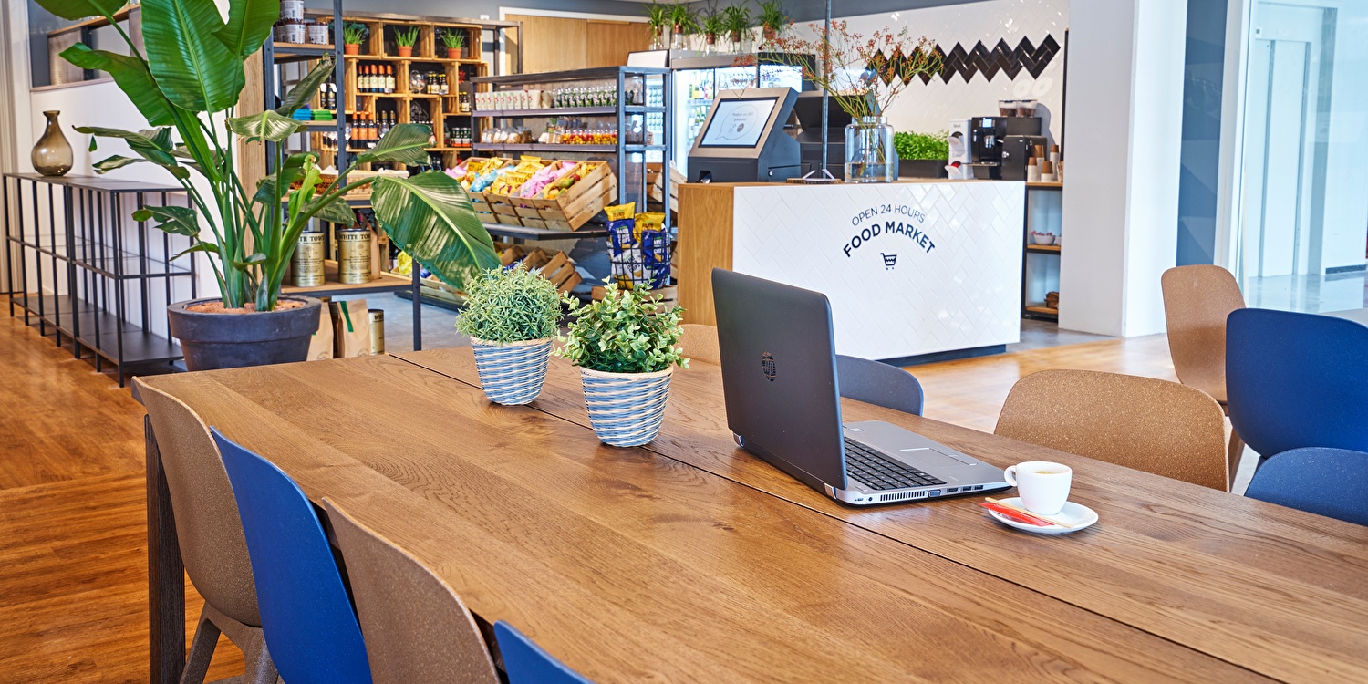 Sfeervolle werk- en ontmoetingsruimte met een lange houten tafel, comfortabele stoelen, groenaccenten en een 24-uurs food market op de achtergrond met snacks, drankjes en versproducten.