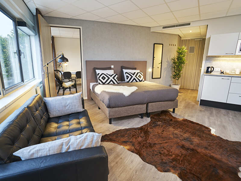 Hotel apartment with modern interior, a double bed, black leather sofa, kitchenette, and a separate area with a desk.